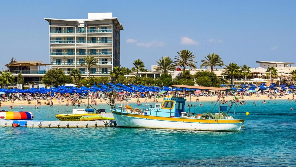 Makronissos strand i Ayia Napa med blå parasoller, båt och turkost vatten.