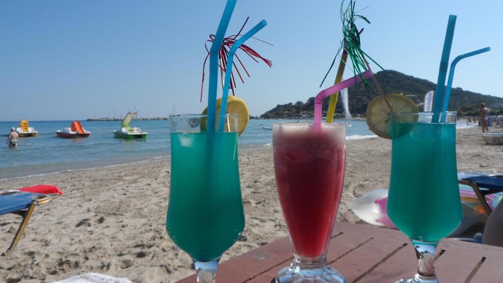 Tre färgglada cocktails på stranden i Zakynthos, Grekland, med sol och hav.