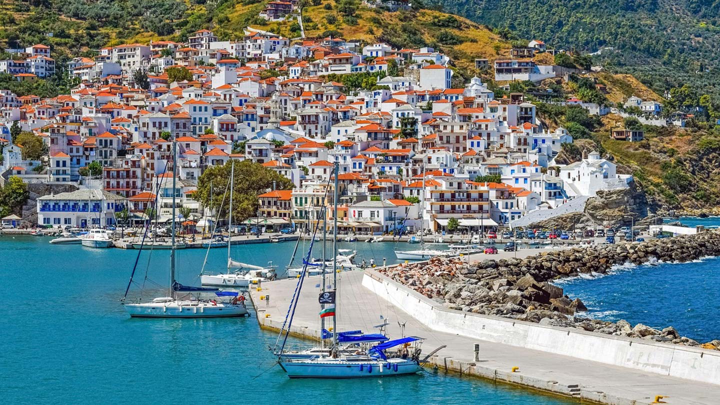 Bilden visar hamnen i Skopelos, Grekland, med vita hus och segelbåtar.