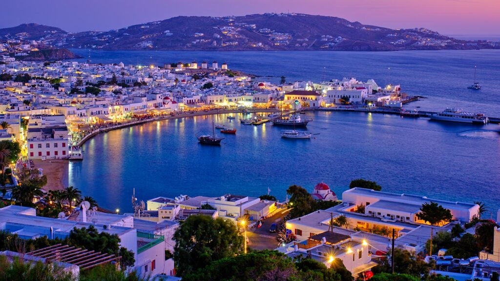 Bilden visar hamnen på Mykonos med båtar och vackra, upplysta byggnader.