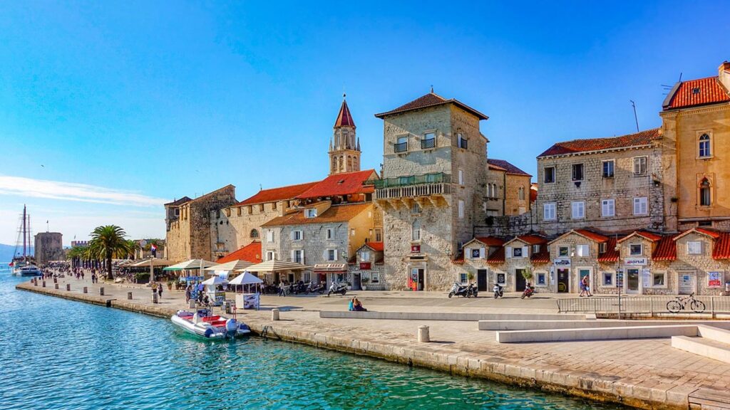 Bilden visar hamnpromenaden i Trogir, Kroatien, med vackra byggnader och turister.