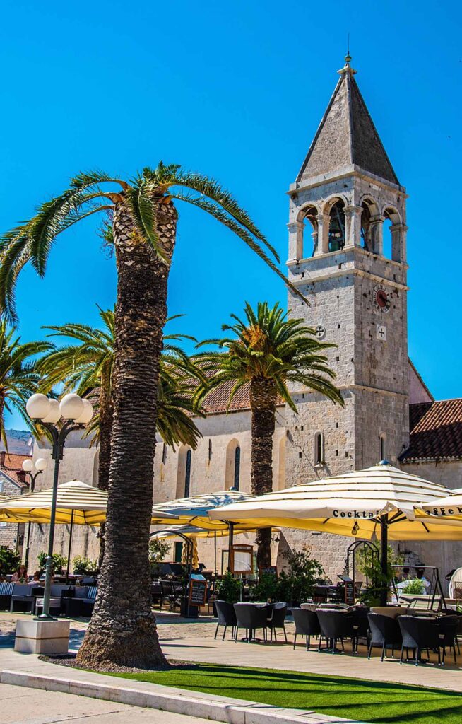 Bild av palmträd och restauranger i Trogir, Kroatien, med en kyrkotorn i bakgrunden.