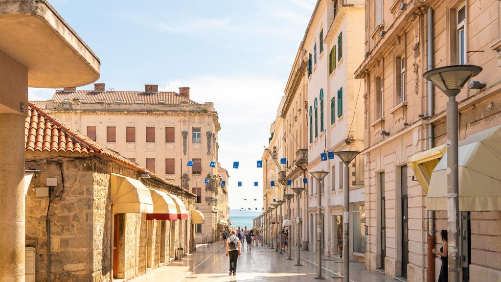 En solbelyst kullerstensgata i Split, Kroatien med människor och hav i bakgrunden.