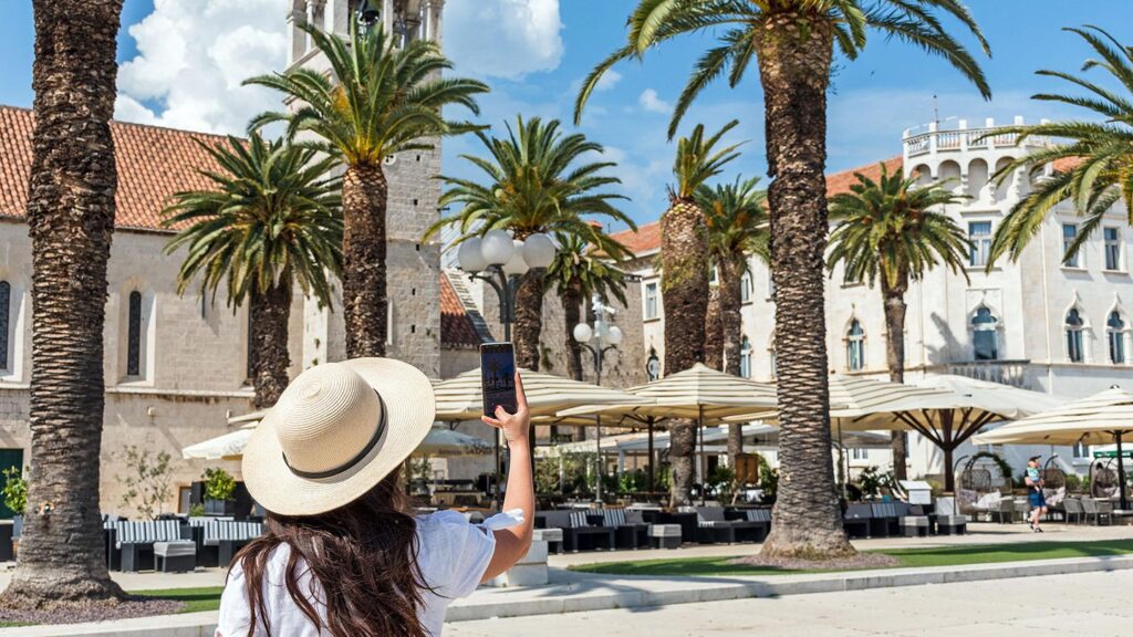 En kvinna tar foto med mobil i Trogir, omgiven av palmträd och byggnader.