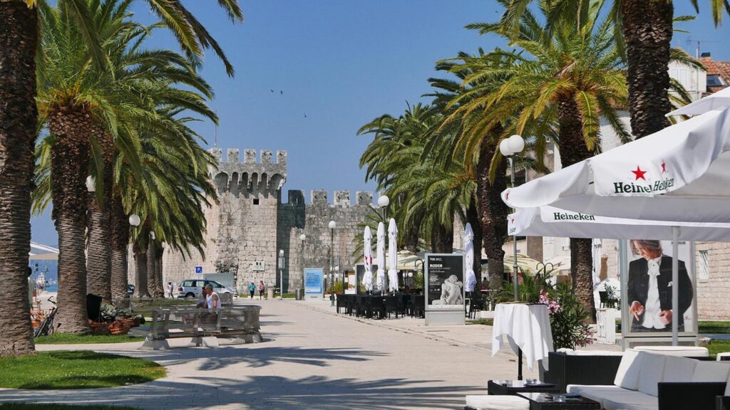 En ljus kustpromenad i Trogir, omgiven av palmträd och historiska byggnader.