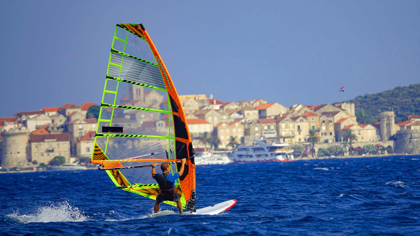 En vindsurfare glider fram på vattnet vid Korčula, Kroatien, med stadens byggnader i bakgrunden.