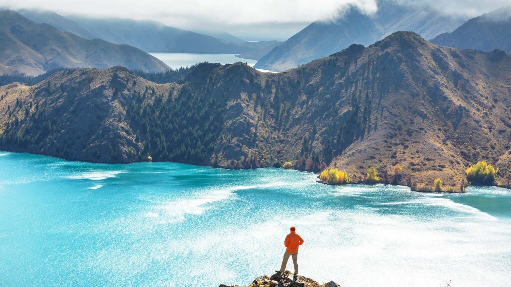 En person står vid en sjö i New Zealand, omgiven av vackra berg.