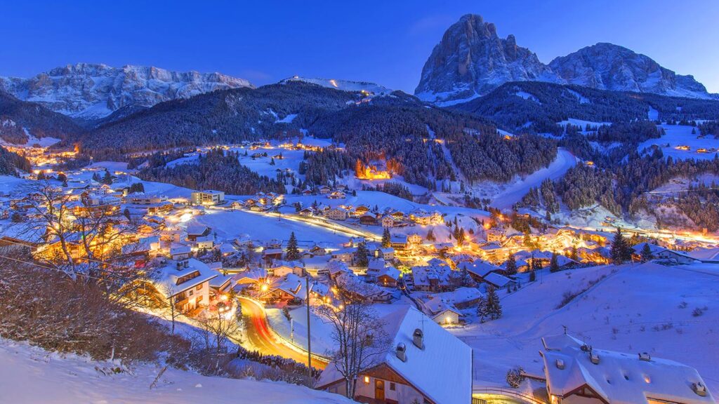 Bilden visar en snötäckt by i Val Gardena, Italien, med fjäll och upplysta hus.