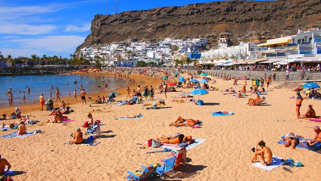 En livlig strand i Puerto de Mogan, Gran Canaria, med solande och badande människor.