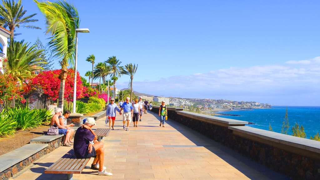 En solig promenad längs stranden i Playa del Inglés, Maspalomas, med palmer och blommor.